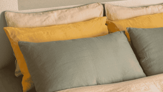 A close-up of a bed styled with multiple pillows, including a prominent sage green pillow in the foreground, a mustard yellow pillow behind it, and cream pillows at the top against a textured wall. 